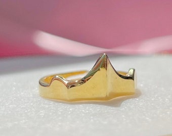 Könnte beinhalten: Ein goldener Ring mit einem Kronendesign. Der Ring liegt auf einer weißen Oberfläche.