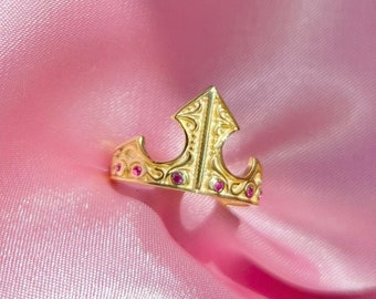 Könnte beinhalten: Ein goldener Kronenring mit roten Edelsteinen. Der Ring liegt auf einem rosa Satin Hintergrund.