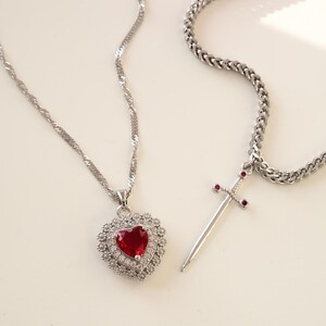 May include: Two silver necklaces with pendants. One necklace has a heart-shaped pendant with a red gemstone surrounded by smaller clear gemstones. The other necklace has a sword pendant with small red gemstones on the hilt.