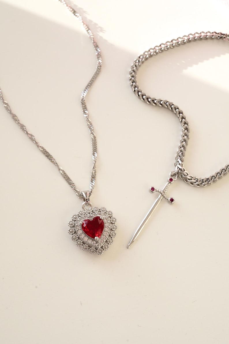 May include: Two silver necklaces with pendants. One necklace has a heart-shaped pendant with a red gemstone surrounded by smaller clear gemstones. The other necklace has a sword pendant with small red gemstones on the hilt.