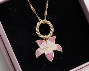 Könnte beinhalten: Eine goldene Kettenkette mit einem rosa und weißen Blumenanhänger. Der Anhänger ist an einem goldenen Ring befestigt.