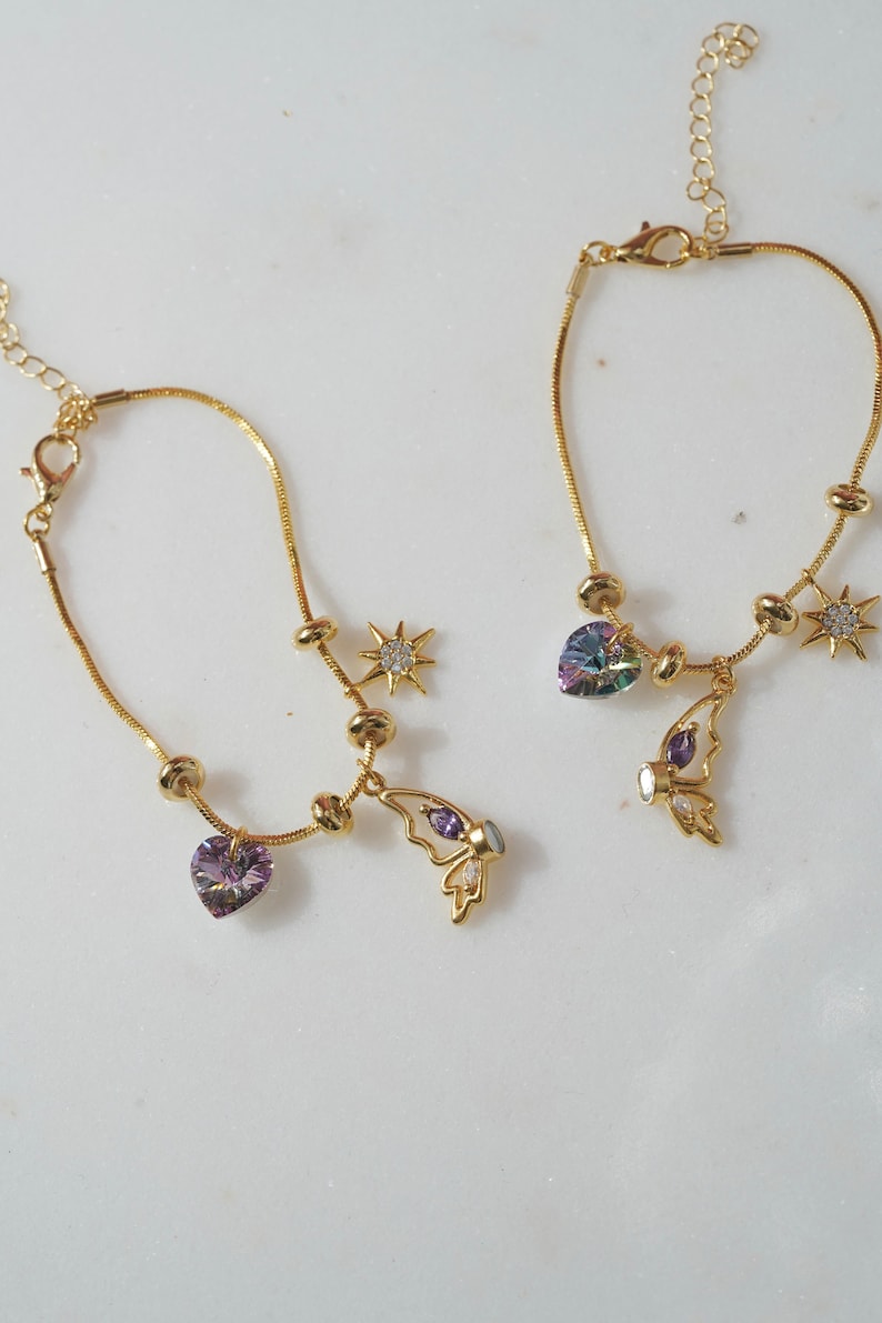 May include: Two gold chain bracelets with charms. One bracelet has a purple heart charm, a butterfly charm, and a star charm. The other bracelet has a rainbow heart charm, a butterfly charm, and a star charm.