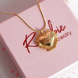 May include: A gold necklace with a heart-shaped pendant surrounded by a halo of smaller hearts. The necklace is displayed in a pink box with the text "Roloie Jewelry" on the front.