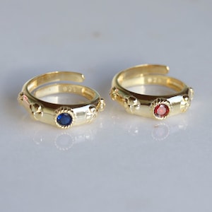 May include: Two gold rings with a blue and red gemstone in the center. Each ring has a decorative design around the gemstone and small decorative accents on the band.