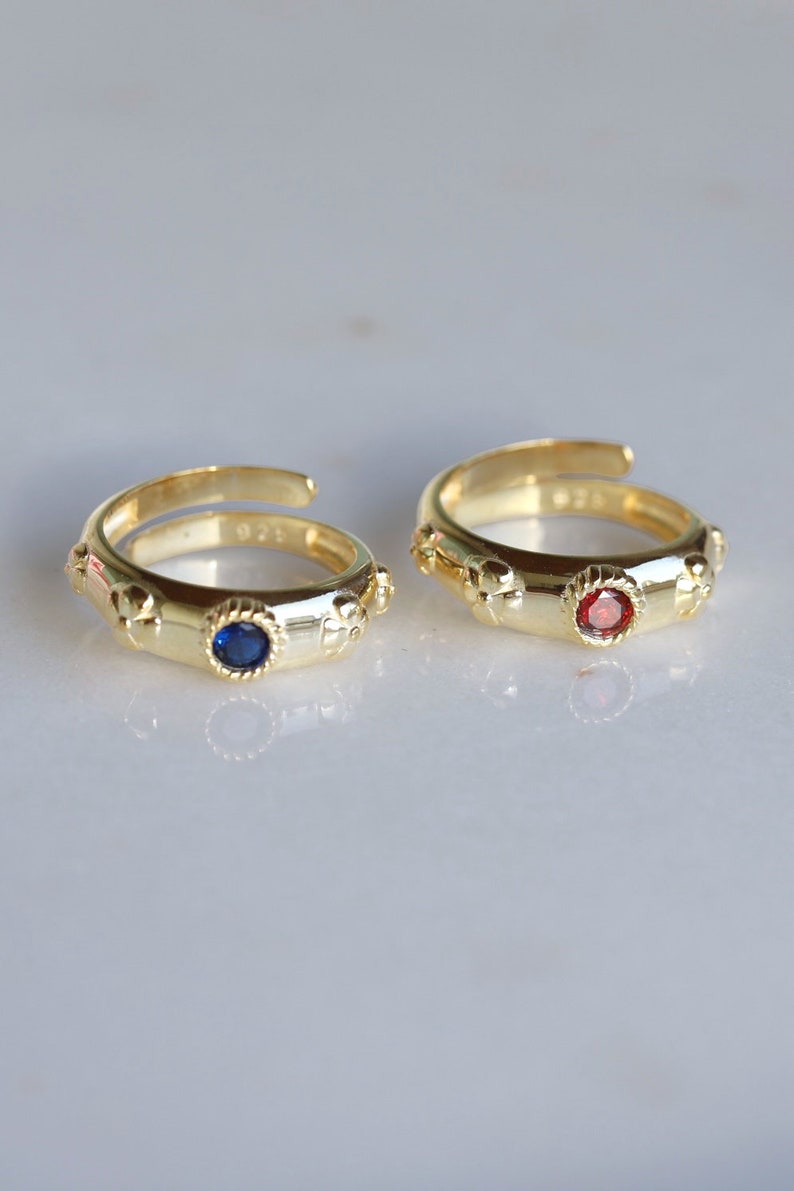 May include: Two gold rings with a blue and red gemstone in the center. Each ring has a decorative design around the gemstone and small decorative accents on the band.