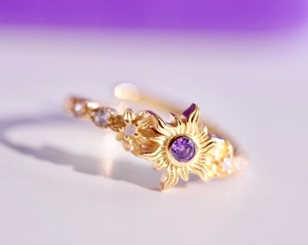 May include: A gold ring with a purple gemstone in the center, surrounded by a sun design. The ring is set with small white gemstones.