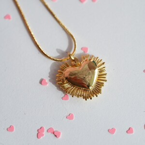 May include: A gold necklace with a heart-shaped pendant surrounded by a sunburst design. The pendant is shiny and reflective. The necklace is on a white surface with pink hearts scattered around it.