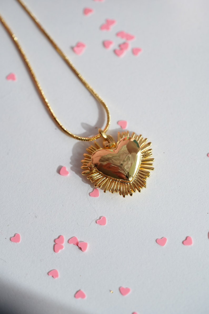 May include: A gold necklace with a heart-shaped pendant surrounded by a sunburst design. The pendant is shiny and reflective. The necklace is on a white surface with pink hearts scattered around it.