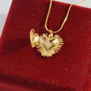 May include: A gold heart-shaped locket necklace with a detailed design. The locket is open and the chain is visible. The necklace is displayed on a red velvet box.