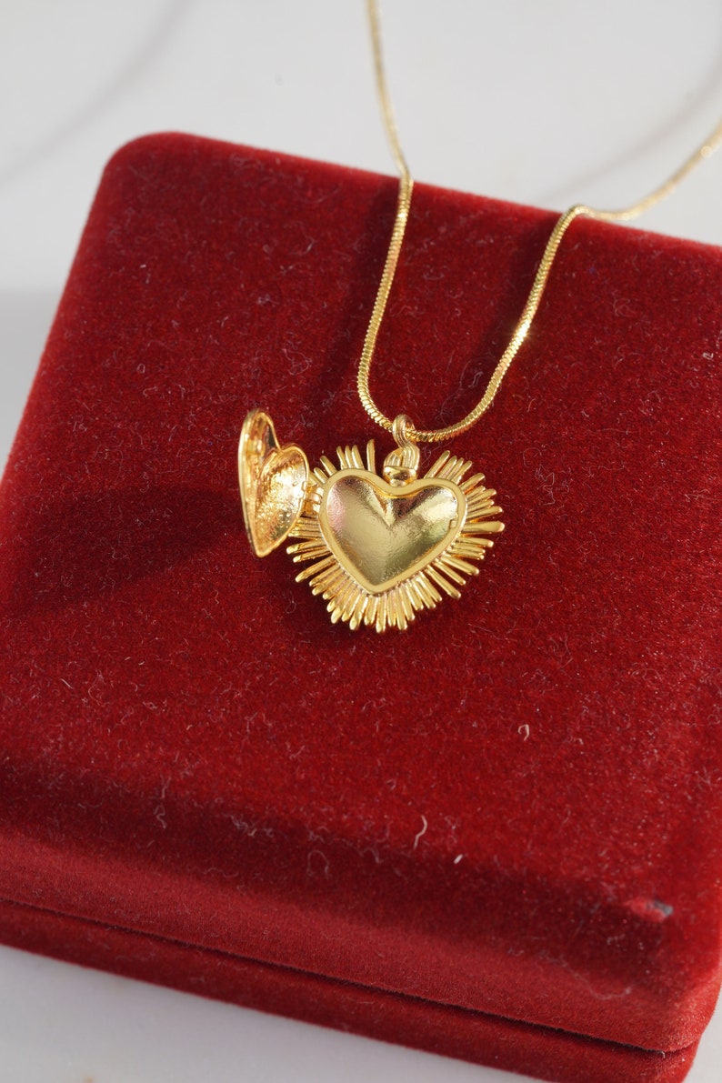 May include: A gold heart-shaped locket necklace with a detailed design. The locket is open and the chain is visible. The necklace is displayed on a red velvet box.