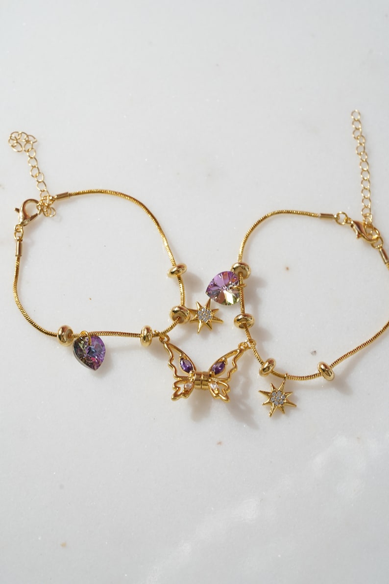 May include: Two gold chain anklets with charms. One anklet has a purple heart charm and a butterfly charm with purple accents. The other anklet has a star charm with clear crystals and a purple heart charm.