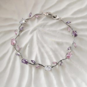 May include: A silver bracelet featuring a vine-like design with oval and round gemstones. The bracelet has pink, purple, and clear stones, resembling leaves and flowers. The clasp is silver and the bracelet is on a white background.