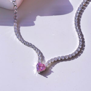 Può includere: Una collana d'argento con un ciondolo a forma di cuore rosa e una fila di pietre di zircone cubico trasparenti.