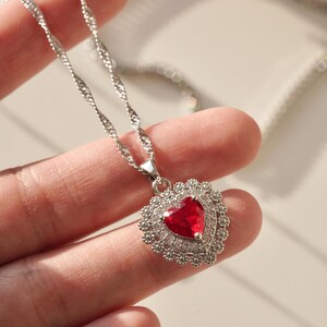 May include: A silver chain necklace with a heart-shaped pendant. The pendant is adorned with small clear stones and features a large red heart-shaped stone in the center.