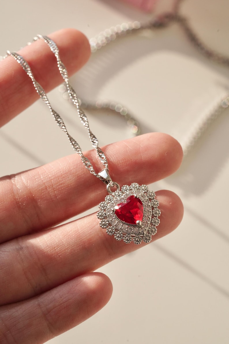 May include: A silver chain necklace with a heart-shaped pendant. The pendant is adorned with small clear stones and features a large red heart-shaped stone in the center.
