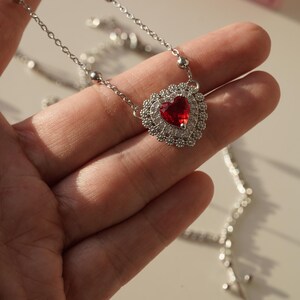 Peut inclure: Un collier en argent avec un pendentif en forme de c&oelig;ur. Le pendentif est orn&eacute; d'une grosse pierre rouge entour&eacute;e de petites pierres claires.