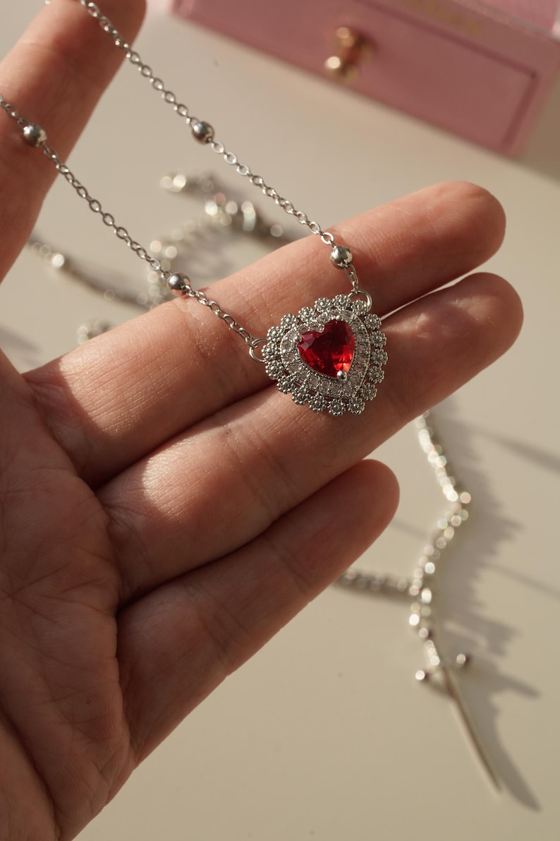 Peut inclure: Un collier en argent avec un pendentif en forme de c&oelig;ur. Le pendentif est orn&eacute; d'une grosse pierre rouge entour&eacute;e de petites pierres claires.