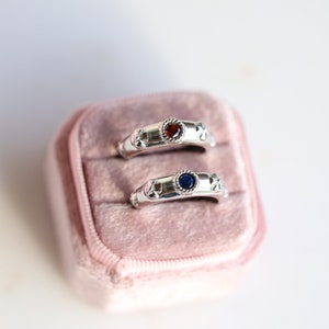 May include: Two silver rings with red and blue gemstones in a pink velvet ring box.