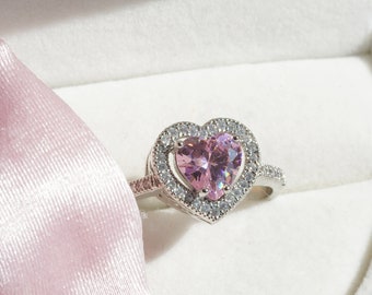 May include: A silver ring with a pink heart-shaped gemstone surrounded by smaller clear gemstones. The ring is in a white heart-shaped box with a pink satin lining.