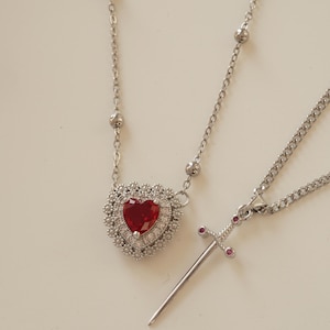 Peut inclure: Deux colliers en argent avec des pendentifs. Un pendentif est de forme de c&oelig;ur avec une pierre pr&eacute;cieuse rouge entour&eacute;e de petites pierres pr&eacute;cieuses claires. L'autre pendentif est une &eacute;p&eacute;e en argent avec de petites pierres pr&eacute;cieuses rouges sur la poign&eacute;e.