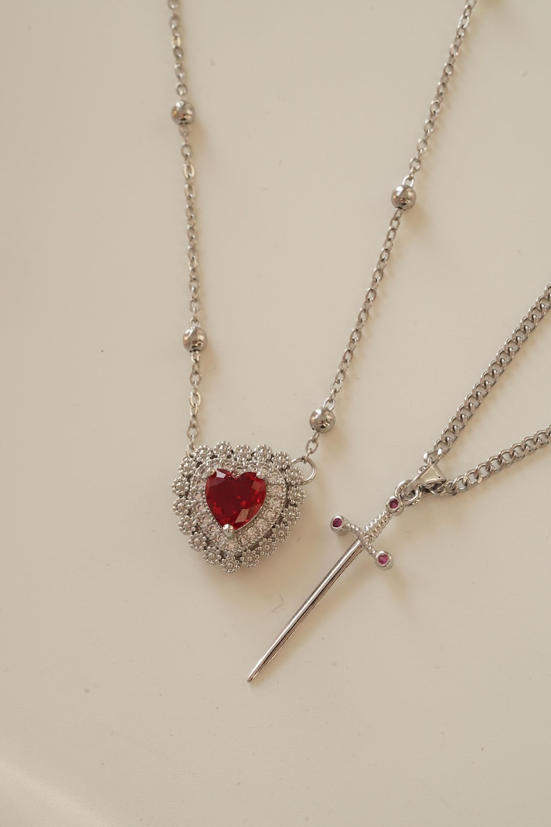 Peut inclure: Deux colliers en argent avec des pendentifs. Un pendentif est de forme de c&oelig;ur avec une pierre pr&eacute;cieuse rouge entour&eacute;e de petites pierres pr&eacute;cieuses claires. L'autre pendentif est une &eacute;p&eacute;e en argent avec de petites pierres pr&eacute;cieuses rouges sur la poign&eacute;e.
