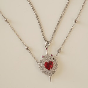 Peut inclure: Un collier en argent avec deux cha&icirc;nes. La cha&icirc;ne la plus longue a un pendentif en forme de c&oelig;ur travers&eacute; par une &eacute;p&eacute;e. Le c&oelig;ur est d&eacute;cor&eacute; de petites pierres blanches et d'une pierre rouge au centre.