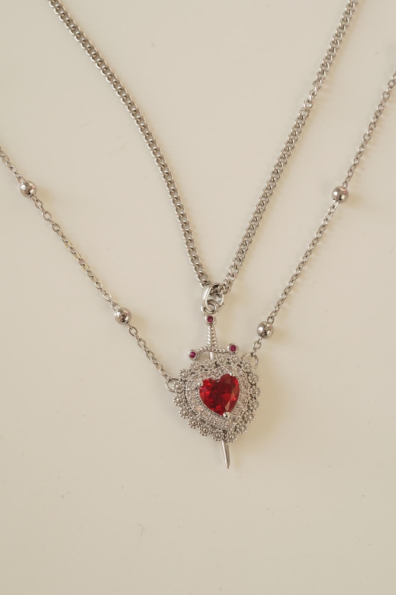 Peut inclure: Un collier en argent avec deux cha&icirc;nes. La cha&icirc;ne la plus longue a un pendentif en forme de c&oelig;ur travers&eacute; par une &eacute;p&eacute;e. Le c&oelig;ur est d&eacute;cor&eacute; de petites pierres blanches et d'une pierre rouge au centre.