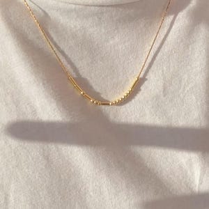 Puede incluir: Un collar de oro con una cadena delicada y una fila de pequeñas cuentas y barras de oro.