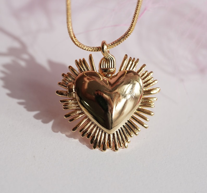 May include: A gold necklace with a heart-shaped pendant. The pendant is surrounded by a halo of rays and has a detailed, textured surface.