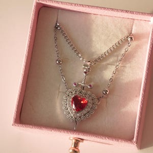 Peut inclure: Un collier en argent avec un pendentif en forme de c&oelig;ur rouge. Le c&oelig;ur est entour&eacute; de petits diamants et d'une &eacute;p&eacute;e en argent.