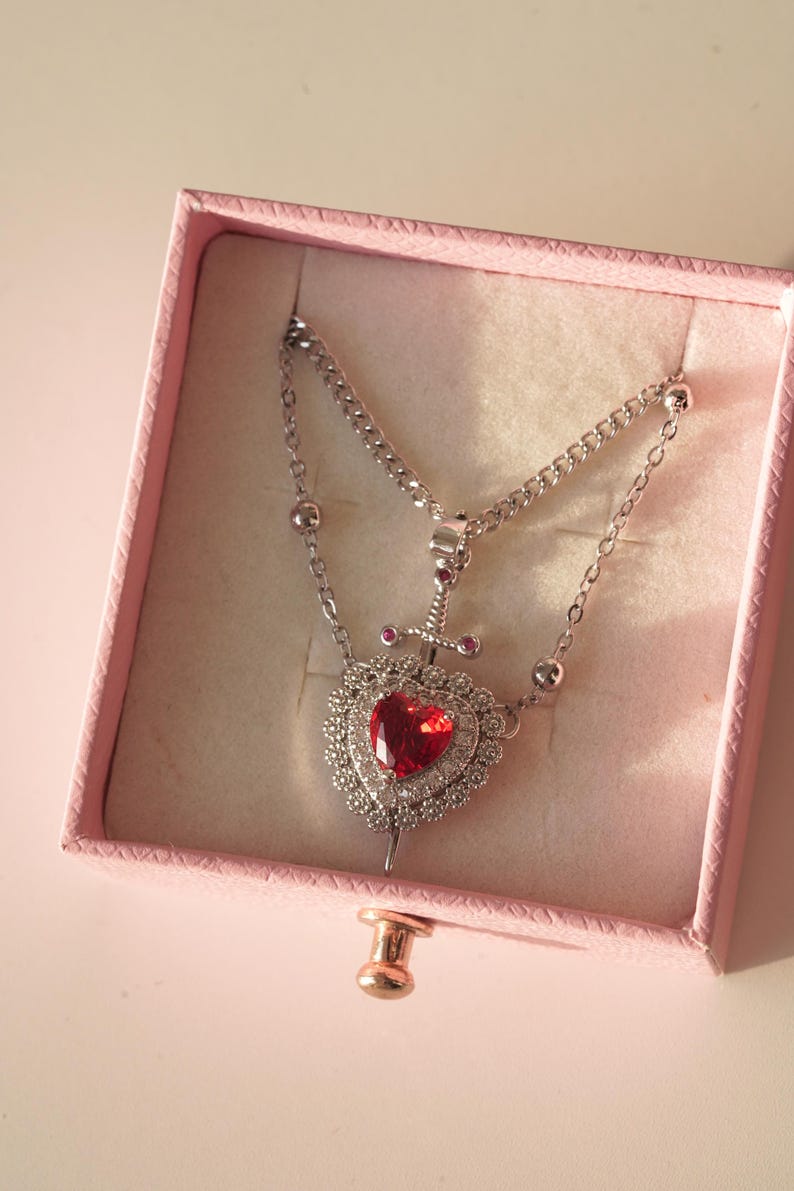 Peut inclure: Un collier en argent avec un pendentif en forme de c&oelig;ur rouge. Le c&oelig;ur est entour&eacute; de petits diamants et d'une &eacute;p&eacute;e en argent.