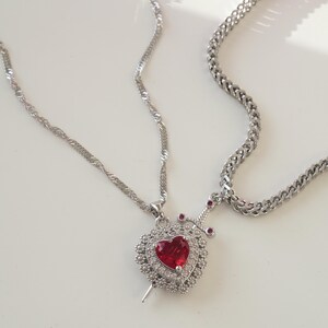 May include: A silver chain necklace with a pendant featuring a red heart with a silver dagger piercing through it. The heart is surrounded by smaller silver stones.