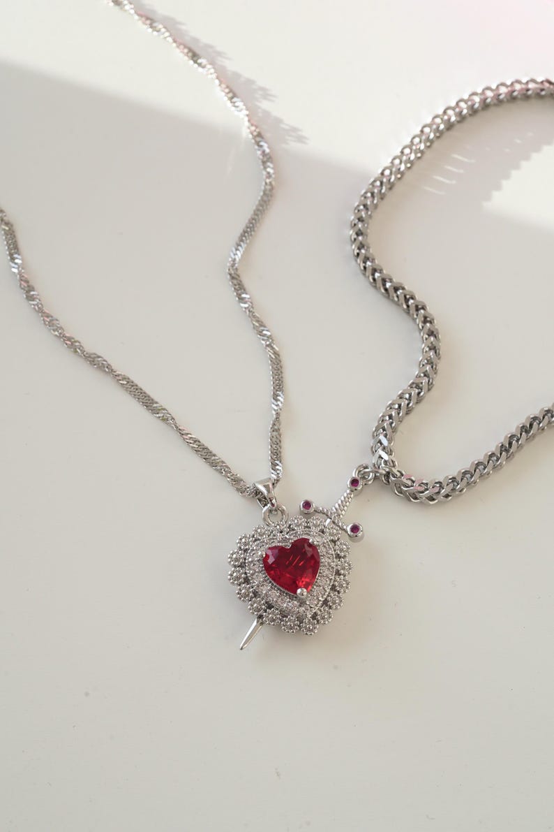 May include: A silver chain necklace with a pendant featuring a red heart with a silver dagger piercing through it. The heart is surrounded by smaller silver stones.