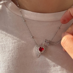 Peut inclure: Un collier en argent avec un pendentif en forme de c&oelig;ur rouge. Le c&oelig;ur est entour&eacute; de petites pierres claires et surmont&eacute; d'une couronne en argent. Le collier est sur une chemise blanche.