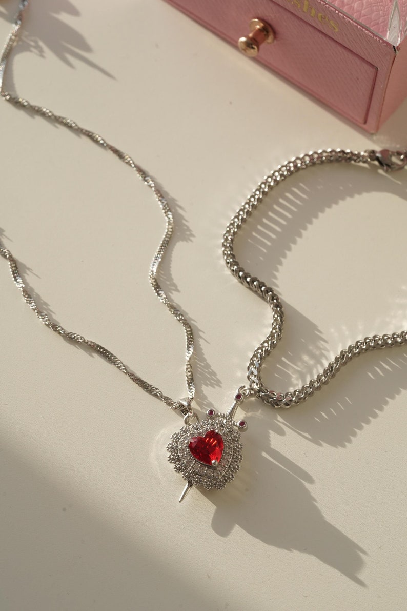 May include: Two silver chains with a pendant featuring a red heart with a crown and a silver sword. The pendant is adorned with small clear crystals.