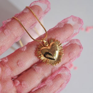 May include: A gold heart pendant necklace with a detailed, textured design. The heart is surrounded by a halo of smaller, pointed shapes. The necklace is displayed on a pink, dotted fabric.
