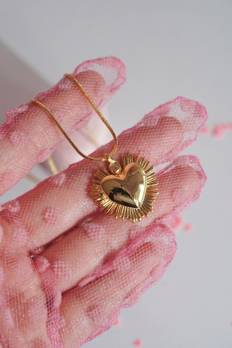 May include: A gold heart pendant necklace with a detailed, textured design. The heart is surrounded by a halo of smaller, pointed shapes. The necklace is displayed on a pink, dotted fabric.