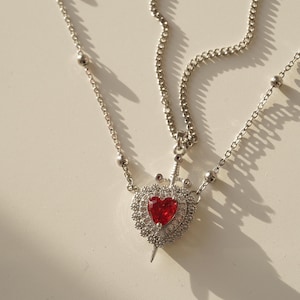 Peut inclure: Un collier en cha&icirc;ne argent avec un pendentif en forme de c&oelig;ur rouge. Le c&oelig;ur est entour&eacute; de petites pierres argent&eacute;es et une &eacute;p&eacute;e argent&eacute;e le traverse au centre.