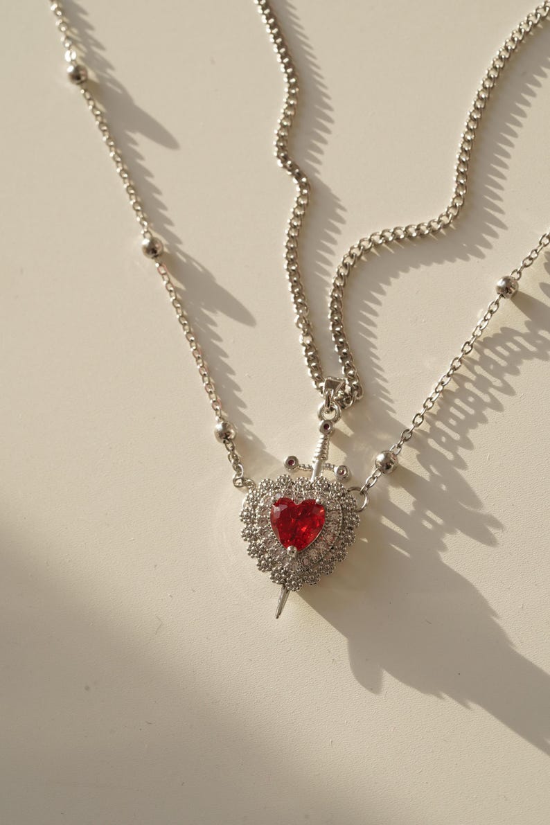 Peut inclure: Un collier en cha&icirc;ne argent avec un pendentif en forme de c&oelig;ur rouge. Le c&oelig;ur est entour&eacute; de petites pierres argent&eacute;es et une &eacute;p&eacute;e argent&eacute;e le traverse au centre.