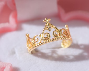May include: A gold crown ring with a small clear stone in the center. The ring is in a pink velvet box.