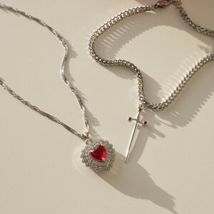 May include: Two silver necklaces with pendants. One necklace has a red heart pendant with clear crystals. The other necklace has a silver sword pendant with red accents.