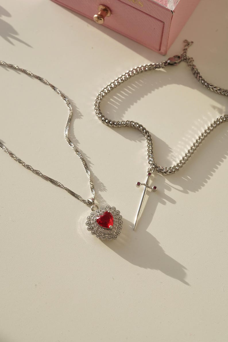 May include: Two silver necklaces with pendants. One necklace has a red heart pendant with clear crystals. The other necklace has a silver sword pendant with red accents.