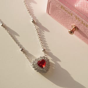 Peut inclure: Un collier en argent avec un pendentif en forme de c&oelig;ur rouge. Le c&oelig;ur est entour&eacute; de petites pierres argent&eacute;es. Le collier est sur une surface blanche.