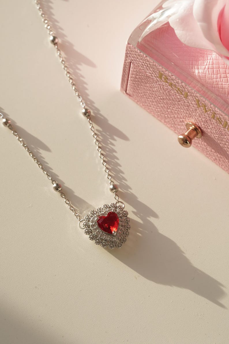 Peut inclure: Un collier en argent avec un pendentif en forme de c&oelig;ur rouge. Le c&oelig;ur est entour&eacute; de petites pierres argent&eacute;es. Le collier est sur une surface blanche.
