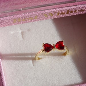 May include: A gold ring with a red bow design. The ring is in a pink box with the words "Best Wishes" on the top.