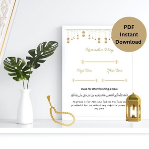 May include: A framed Ramadan Day print with gold accents, featuring text in English and Arabic. The print includes sections for Fajr and Iftar times, and a prayer. A white vase with green leaves, a gold lantern, and a gold beaded chain are also visible.