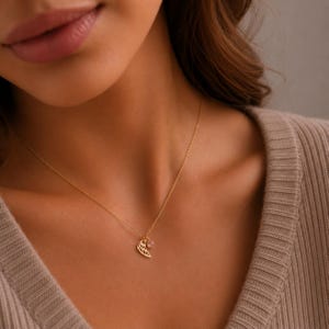 May include: A delicate gold necklace featuring a wing pendant and a small, round gemstone. The necklace is displayed on a person wearing a beige sweater. The wing pendant has intricate detailing.