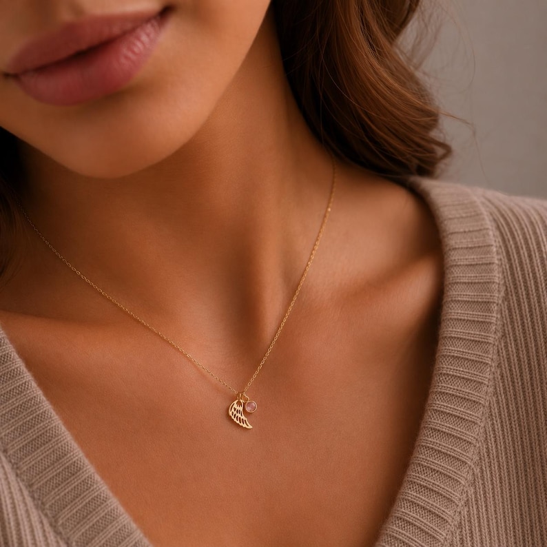 May include: A delicate gold necklace featuring a wing pendant and a small, round gemstone. The necklace is displayed on a person wearing a beige sweater. The wing pendant has intricate detailing.