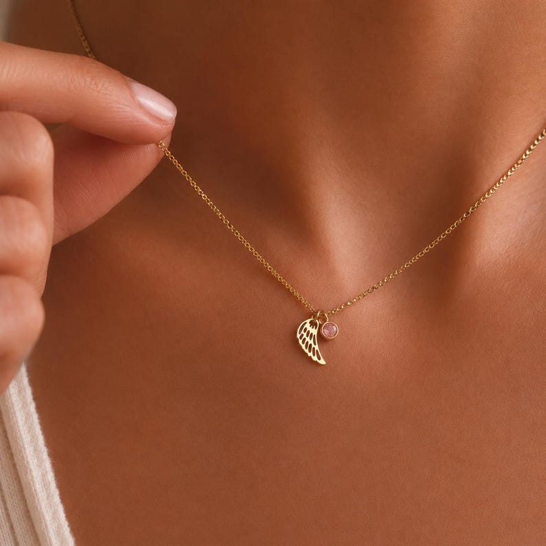 May include: A gold-colored necklace with a delicate wing pendant and a small pink gemstone charm. The necklace is displayed on a person's neck, showcasing the jewelry's design and color. The chain is thin and the wing pendant is detailed.