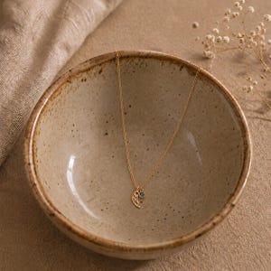 May include: A delicate gold necklace with a small wing pendant and a blue gemstone. The necklace is displayed in a speckled ceramic bowl, with a neutral-toned background and dried flowers.
